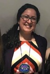 headshot of a smiling woman with black hair tied to the side. she is wearing dangly earrings and a long necklace, as well as a diamond patterned top and black cardigan