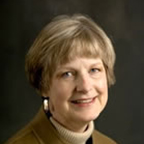 headshot of a smiling woman with light, short hair. she is wearing earrings, beige turtleneck, and a brown blazer