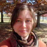 Selfie of a woman with tied back hair. She is wearing a scarf and sweater while standing outside with trees in the background