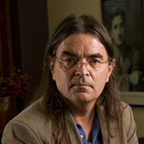headshot of a man with long, brown hair and glasses. he is wearing a blue button-up and tan blazer